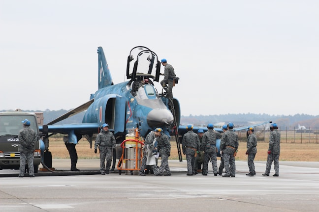 A group of military personnel in uniform are gathered around a fighter jet on an airfield. Some personnel are on the ground, while one person is on top of the aircraft. The jet is a dark blue color with various markings. A vehicle is parked nearby, and the area has an open, expansive feel with a distant tree line.