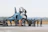 A group of military personnel in uniform are gathered around a fighter jet on an airfield. Some personnel are on the ground, while one person is on top of the aircraft. The jet is a dark blue color with various markings. A vehicle is parked nearby, and the area has an open, expansive feel with a distant tree line.