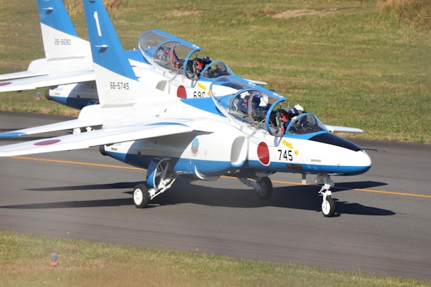Two blue and white jet aircraft are positioned on a runway, with grassy areas surrounding the tarmac. The planes are marked with numbers and insignia, and pilots are visible inside the cockpit, readying for takeoff.