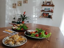 The image features a wooden table set with plates of fresh salad and a potato dish with cheese and scrambled eggs. In the background, there is a plant in a pot, photo frames, and decorative items on shelves attached to the wall. The room has a cozy, homely atmosphere with natural light streaming through, casting soft shadows.