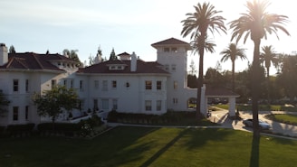 A large, elegant white mansion with a red-tiled roof, surrounded by lush greenery and tall palm trees. The bright sunlight casts long shadows and illuminates the serene setting.