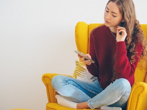 A young woman with long, curly hair is sitting cross-legged on a bright yellow armchair. She is wearing a red sweater and blue jeans, holding a smartphone in one hand while the other hand is touching her chin in a thoughtful manner.