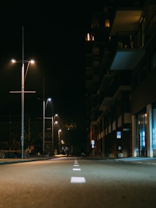A quiet street at night illuminated by rows of streetlights. The left side features tall poles with modern light fixtures casting a warm glow. An apartment building runs along the right side, with lit windows and balconies visible. A lone cyclist rides down the middle of the deserted road, adding a sense of solitude to the scene.