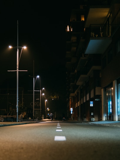 A quiet street at night illuminated by rows of streetlights. The left side features tall poles with modern light fixtures casting a warm glow. An apartment building runs along the right side, with lit windows and balconies visible. A lone cyclist rides down the middle of the deserted road, adding a sense of solitude to the scene.