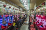 A brightly lit gaming arcade filled with rows of colorful pachinko machines. People are seated at various machines, engaged in playing. Red and blue neon lights dominate the room, with advertisements and signs displayed above the machines. The ceiling is lined with fluorescent lights, reflecting off the glossy floor.