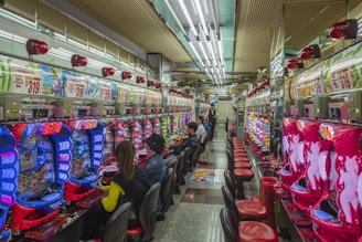 A brightly lit gaming arcade filled with rows of colorful pachinko machines. People are seated at various machines, engaged in playing. Red and blue neon lights dominate the room, with advertisements and signs displayed above the machines. The ceiling is lined with fluorescent lights, reflecting off the glossy floor.