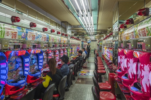 A brightly lit gaming arcade filled with rows of colorful pachinko machines. People are seated at various machines, engaged in playing. Red and blue neon lights dominate the room, with advertisements and signs displayed above the machines. The ceiling is lined with fluorescent lights, reflecting off the glossy floor.