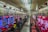 A brightly lit gaming arcade filled with rows of colorful pachinko machines. People are seated at various machines, engaged in playing. Red and blue neon lights dominate the room, with advertisements and signs displayed above the machines. The ceiling is lined with fluorescent lights, reflecting off the glossy floor.