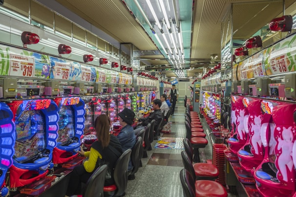 A brightly lit gaming arcade filled with rows of colorful pachinko machines. People are seated at various machines, engaged in playing. Red and blue neon lights dominate the room, with advertisements and signs displayed above the machines. The ceiling is lined with fluorescent lights, reflecting off the glossy floor.