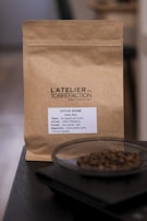 A brown paper coffee bag labeled 'L'ATELIER de TORRÉFACTION' with details about the coffee including region, altitude, and tasting notes. In front of the bag, there is a dish containing coffee beans placed on a dark surface.