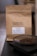 A brown paper coffee bag labeled 'L'ATELIER de TORRÉFACTION' with details about the coffee including region, altitude, and tasting notes. In front of the bag, there is a dish containing coffee beans placed on a dark surface.