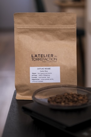 A brown paper coffee bag labeled 'L'ATELIER de TORR&Eacute;FACTION' with details about the coffee including region, altitude, and tasting notes. In front of the bag, there is a dish containing coffee beans placed on a dark surface.