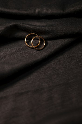 Two gold rings rest on a textured, dark fabric with gentle folds. The lighting highlights the metallic sheen of the rings against the backdrop.