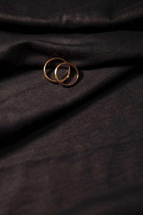 Two gold rings rest on a textured, dark fabric with gentle folds. The lighting highlights the metallic sheen of the rings against the backdrop.