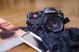 A vintage Leica camera is placed on a wooden table along with a book featuring a partially visible cover image. There is also a smartphone lying next to the camera, partially obstructed by a dark cloth. The scene suggests a casual, creative setting.