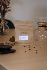 A minimalist coffee setup featuring a brown paper bag of coffee labeled 'L'Atelier de Torréfaction' placed on a wooden table. Scattered coffee beans are visible around the bag. An empty Chemex coffee maker and a glass container with coffee beans are also present, along with a vase containing dried flowers and a fallen leaf, adding a rustic touch.