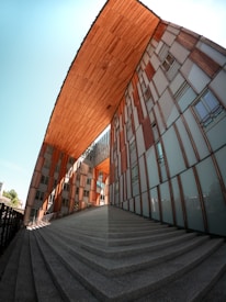 A modern architectural structure with a distinctive geometric design. The building features a large wooden overhang and glass-and-wood facade panels. Wide concrete steps lead up towards the main entrance. The clear blue sky provides a striking backdrop, enhancing the structure's contemporary aesthetic.