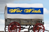 A classic covered wagon with a white canvas top and blue and yellow text reading 'West Winds.' The wagon has red wheels and a wooden structure underneath the canvas. The wagon is set against a partly cloudy sky.