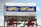 A classic covered wagon with a white canvas top and blue and yellow text reading 'West Winds.' The wagon has red wheels and a wooden structure underneath the canvas. The wagon is set against a partly cloudy sky.