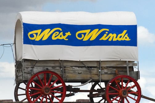 A classic covered wagon with a white canvas top and blue and yellow text reading 'West Winds.' The wagon has red wheels and a wooden structure underneath the canvas. The wagon is set against a partly cloudy sky.