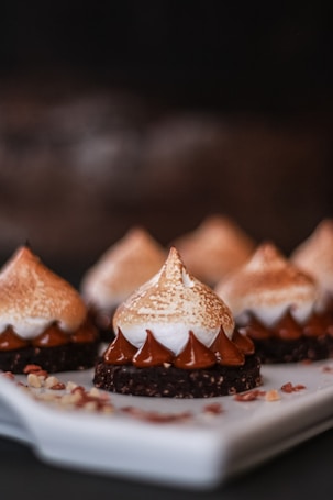 Delicate pastries with a chocolate base, topped with swirls of chocolate ganache and a lightly toasted meringue. The pastries are artfully arranged on a white platter with small nut pieces scattered around.