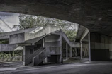 An architectural scene featuring a concrete structure with geometric lines and shadows. The structure includes a series of stairs, platforms, and columns, with elements creating an interplay of light and shadow. Trees and greenery are visible in the background, lending a natural contrast to the stark gray of the concrete.