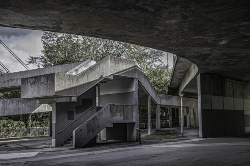 An architectural scene featuring a concrete structure with geometric lines and shadows. The structure includes a series of stairs, platforms, and columns, with elements creating an interplay of light and shadow. Trees and greenery are visible in the background, lending a natural contrast to the stark gray of the concrete.