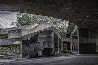 An architectural scene featuring a concrete structure with geometric lines and shadows. The structure includes a series of stairs, platforms, and columns, with elements creating an interplay of light and shadow. Trees and greenery are visible in the background, lending a natural contrast to the stark gray of the concrete.