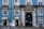 A blue building with white window frames and a large black door that has 'Villa Sousa' and '1890' written on it. It features balconies with black wrought iron railings, potted plants, and a couple of small shops at ground level, including a bookstore and a small convenience store.