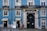 A blue building with white window frames and a large black door that has 'Villa Sousa' and '1890' written on it. It features balconies with black wrought iron railings, potted plants, and a couple of small shops at ground level, including a bookstore and a small convenience store.