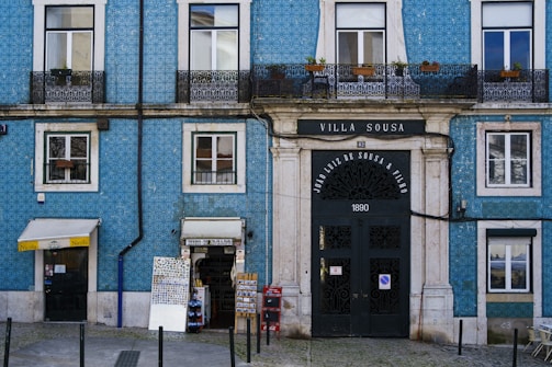 A blue building with white window frames and a large black door that has 'Villa Sousa' and '1890' written on it. It features balconies with black wrought iron railings, potted plants, and a couple of small shops at ground level, including a bookstore and a small convenience store.