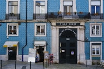 A blue building with white window frames and a large black door that has 'Villa Sousa' and '1890' written on it. It features balconies with black wrought iron railings, potted plants, and a couple of small shops at ground level, including a bookstore and a small convenience store.
