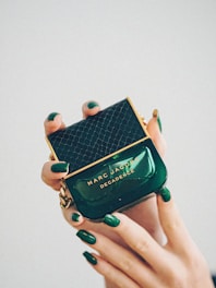 A luxurious perfume bottle being held delicately by hands with matching dark green nail polish. The bottle is green with gold accents and features a textured pattern on the cap. The brand name 'Marc Jacobs' and the perfume name 'Decadence' are prominently displayed.
