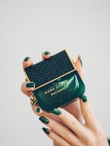 A luxurious perfume bottle being held delicately by hands with matching dark green nail polish. The bottle is green with gold accents and features a textured pattern on the cap. The brand name 'Marc Jacobs' and the perfume name 'Decadence' are prominently displayed.