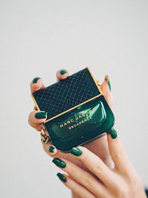 A luxurious perfume bottle being held delicately by hands with matching dark green nail polish. The bottle is green with gold accents and features a textured pattern on the cap. The brand name 'Marc Jacobs' and the perfume name 'Decadence' are prominently displayed.