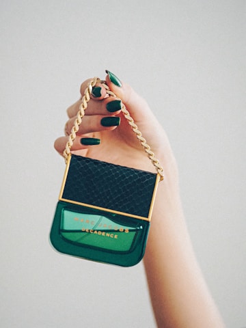 A hand with polished green nails holds a Marc Jacobs Decadence perfume bottle designed to look like a handbag with a golden chain handle and a black textured cap.