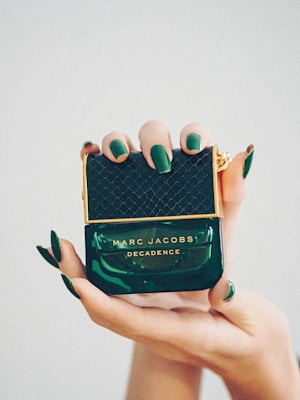 Hands with green nails hold a luxurious bottle of Marc Jacobs Decadence perfume, characterized by its deep green color and gold accents.