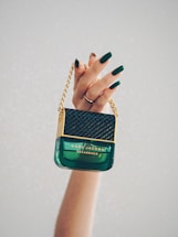 A hand with dark green painted nails holds a luxurious Marc Jacobs Decadence perfume bottle, which features a distinctive handbag design with gold chain detailing.