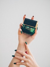 Two hands with elegant green nail polish gracefully hold a perfume bottle. The bottle is dark green with a textured black cap and gold accents, featuring the label 'Marc Jacobs Decadence.'