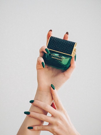 Two hands with elegant green nail polish gracefully hold a perfume bottle. The bottle is dark green with a textured black cap and gold accents, featuring the label 'Marc Jacobs Decadence.'