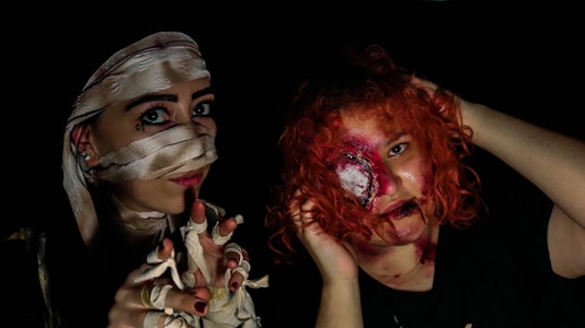 Two individuals in costume. One person is dressed as a mummy with fabric wrappings covering parts of their face and hands. The other person has a special effects makeup, with a gruesome, bloody appearance around one eye and vibrant red curly hair.