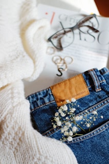 A close-up view featuring a pair of blue jeans with a leather patch, decorated with small white flowers tucked into the pocket. Next to it is a cozy knitted cream-colored sweater, a magazine, and a pair of stylish glasses on top.