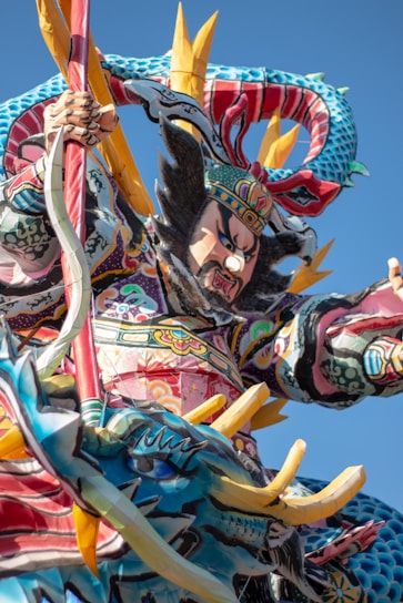 A vibrant and colorful sculpture featuring a fierce warrior figure adorned with intricate patterns and traditional attire. The warrior holds a weapon and is accompanied by a majestic dragon, both elements showcasing elaborate designs. The background is a clear blue sky, enhancing the vivid colors of the sculpture.