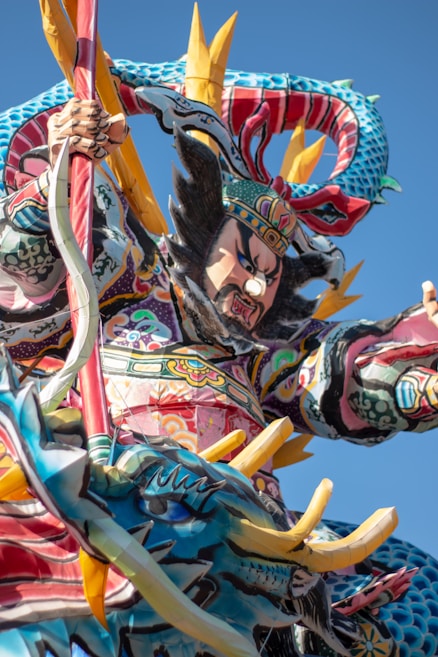 A vibrant and colorful sculpture featuring a fierce warrior figure adorned with intricate patterns and traditional attire. The warrior holds a weapon and is accompanied by a majestic dragon, both elements showcasing elaborate designs. The background is a clear blue sky, enhancing the vivid colors of the sculpture.