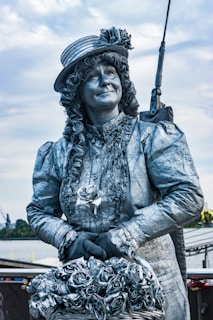A person is dressed as a silver-painted statue, wearing elaborate clothing that includes a hat with flowers and lace-trimmed sleeves. They are holding a basket of silver roses and are made up to look like a living statue, with detailed facial features and a composed expression.