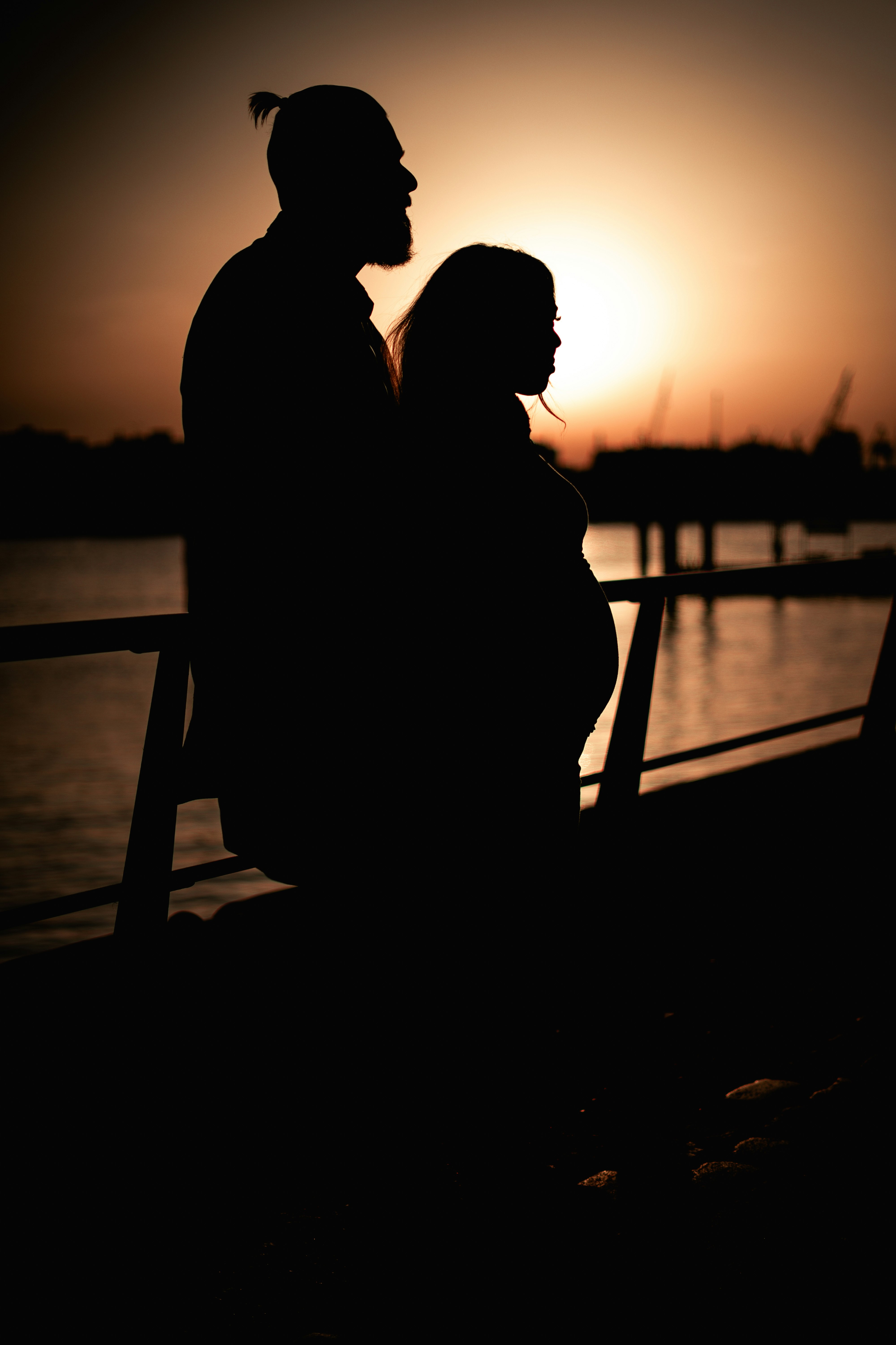 silhouette of man and woman standing beside body of water during sunset