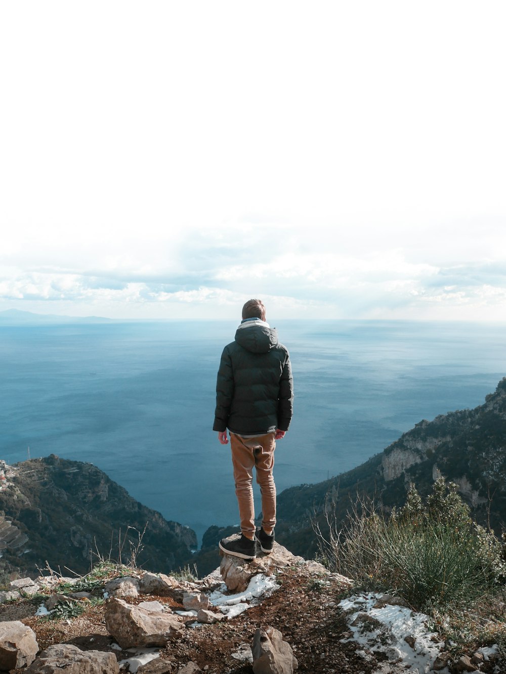 昼間、茶色の岩層の上に立って海を眺める黒いジャケットの男