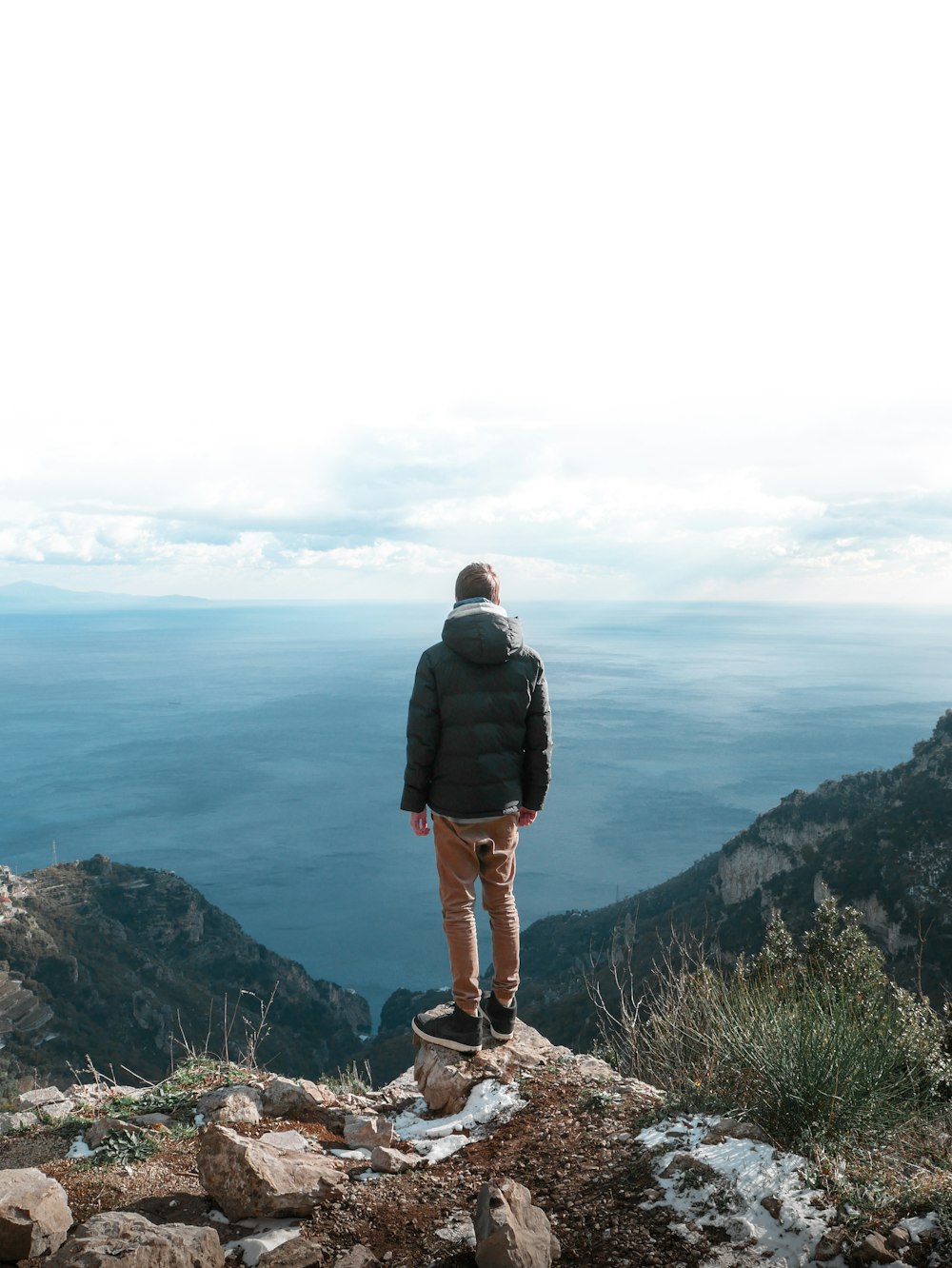 man in black jacket standing on brown rock formation looking at the sea during daytime