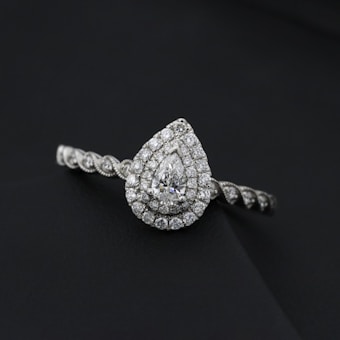 A pear-shaped diamond ring with a detailed band, prominently set against a dark background. The diamond is surrounded by smaller, intricately arranged gems.