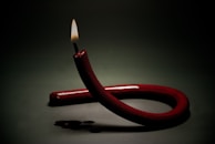 A unique candle with a deep red color is twisted into a loop, with its wick lit and burning steadily. The background is dark, enhancing the candle's bright flame and glossy surface. Wax drips can be seen on the surface where the candle rests.